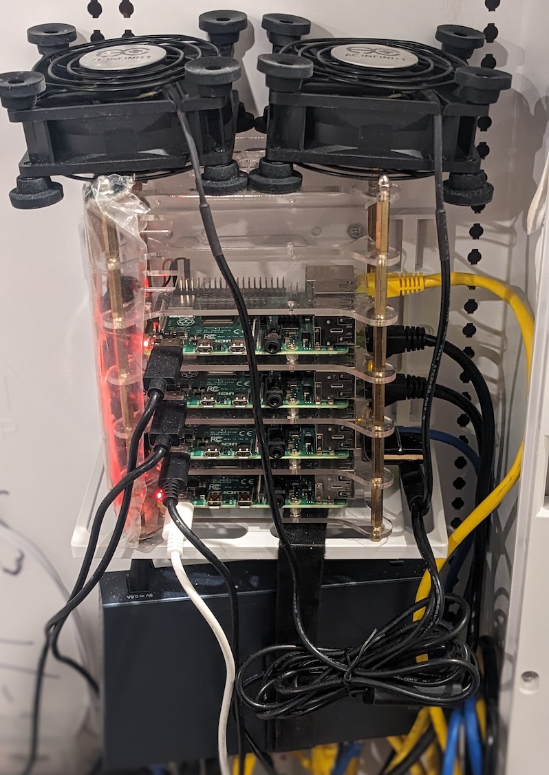 Four Raspberry Pi 4 boards stacked in a clear acrylic case, two cooling fans mounted on top, a network switch and power supply below, with ethernet and USB-C cables running between them.